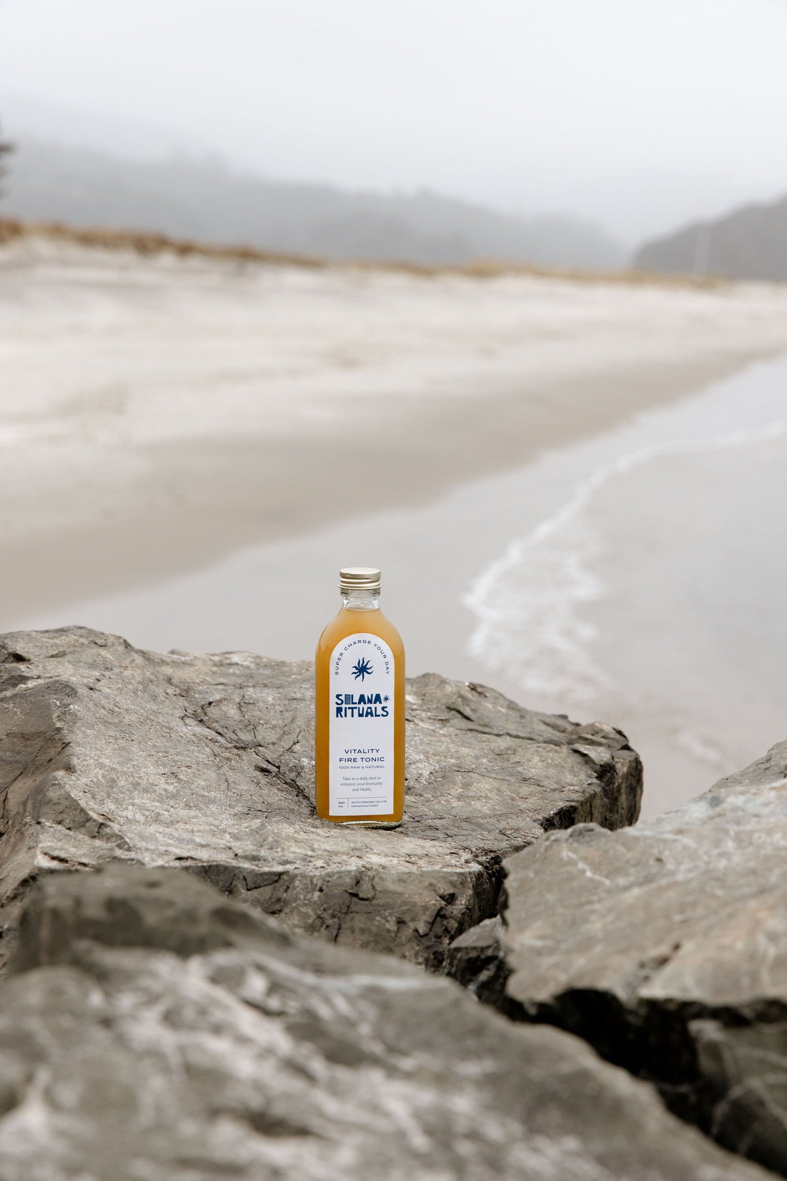 A bottle of Silana Rituals Vitality Fire Tonic resting on a rock at the beach with misty mountains and an overcast sky in the background.